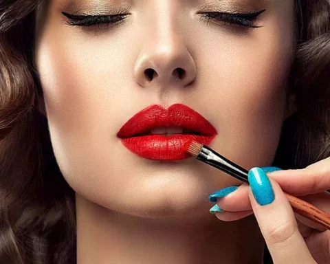 Primer plano de una mujer con sombra de ojos dorada mientras una mano con las uñas pintadas de azul le aplica pintalabios rojo con una brocha de maquillaje.