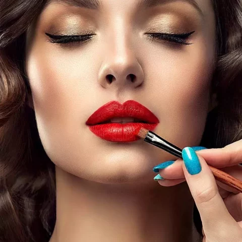 Close-up of a woman with gold eyeshadow as red lipstick is being applied with a makeup brush by a hand with blue nails.
