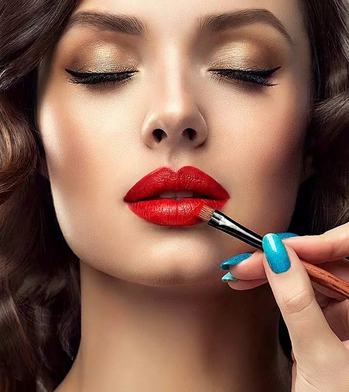 Close-up of a woman with gold eyeshadow as red lipstick is being applied with a makeup brush by a hand with blue nails.