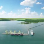 Aerial view of an offshore port with yellow gantry cranes on floating platforms and a docked cargo ship connected by a pier.