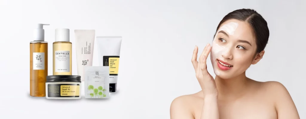 Smiling woman applying cream to her face beside a row of skincare products on a white surface.