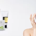 Smiling woman applying cream to her face beside a row of skincare products on a white surface.