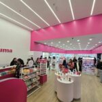 Bright cosmetics store interior with pink walls, white counters, circular display plinths, and customers browsing makeup products.