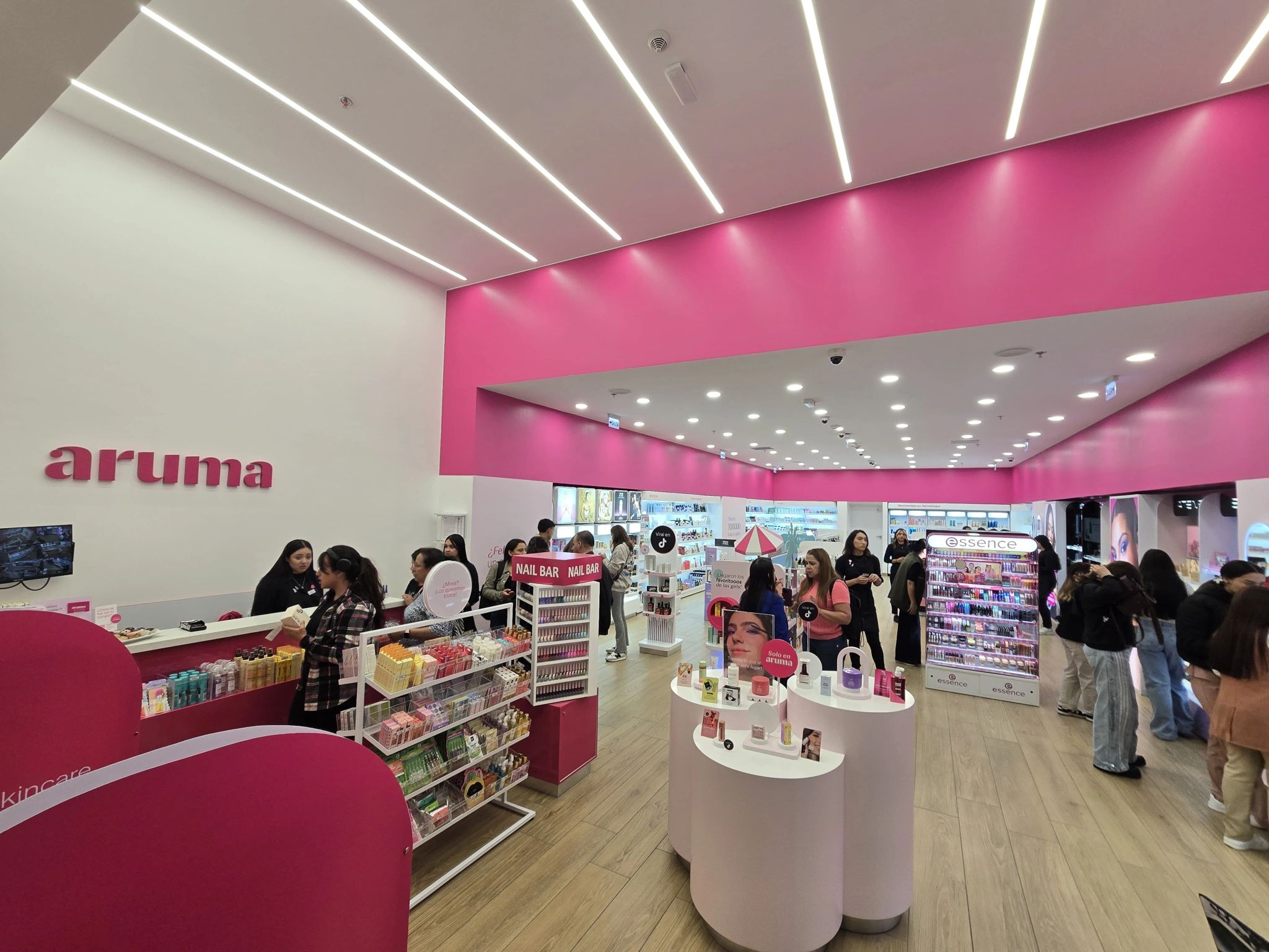 Bright cosmetics store interior with pink walls, white counters, circular display plinths, and customers browsing makeup products.