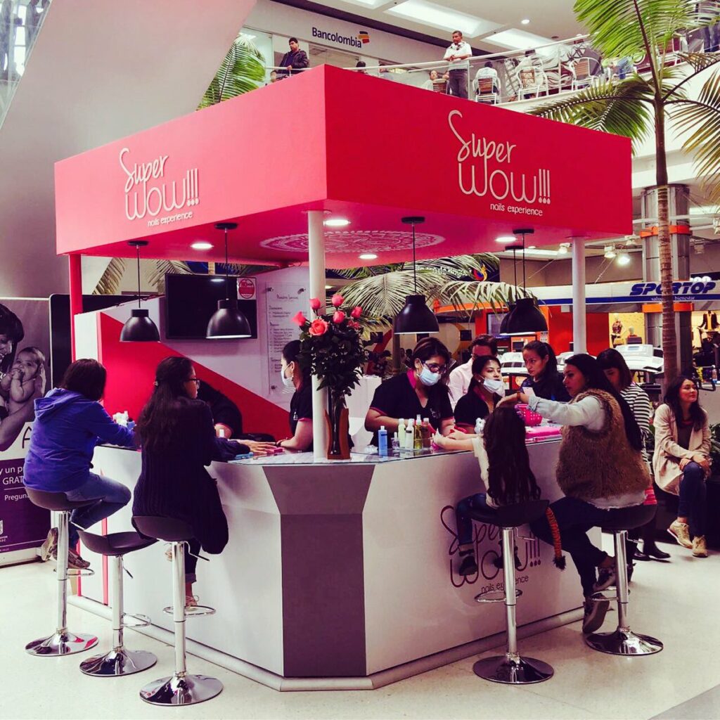 Pink nail-salon kiosk with a bright canopy labeled 'Super WOW!!! nails experience' at a mall, staff and customers seated around a curved counter.
