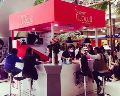 Pink nail-salon kiosk with a bright canopy labeled 'Super WOW!!! nails experience' at a mall, staff and customers seated around a curved counter.