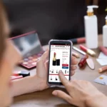 Person holding a smartphone showing an online lipstick product page amid makeup items on a table.