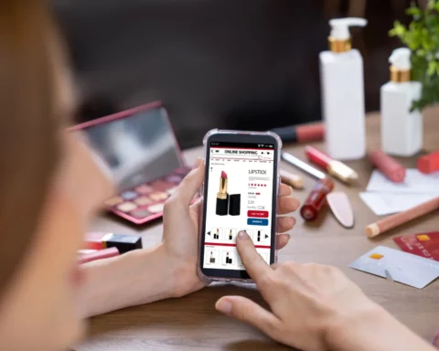 Person holding a smartphone showing an online lipstick product page amid makeup items on a table.