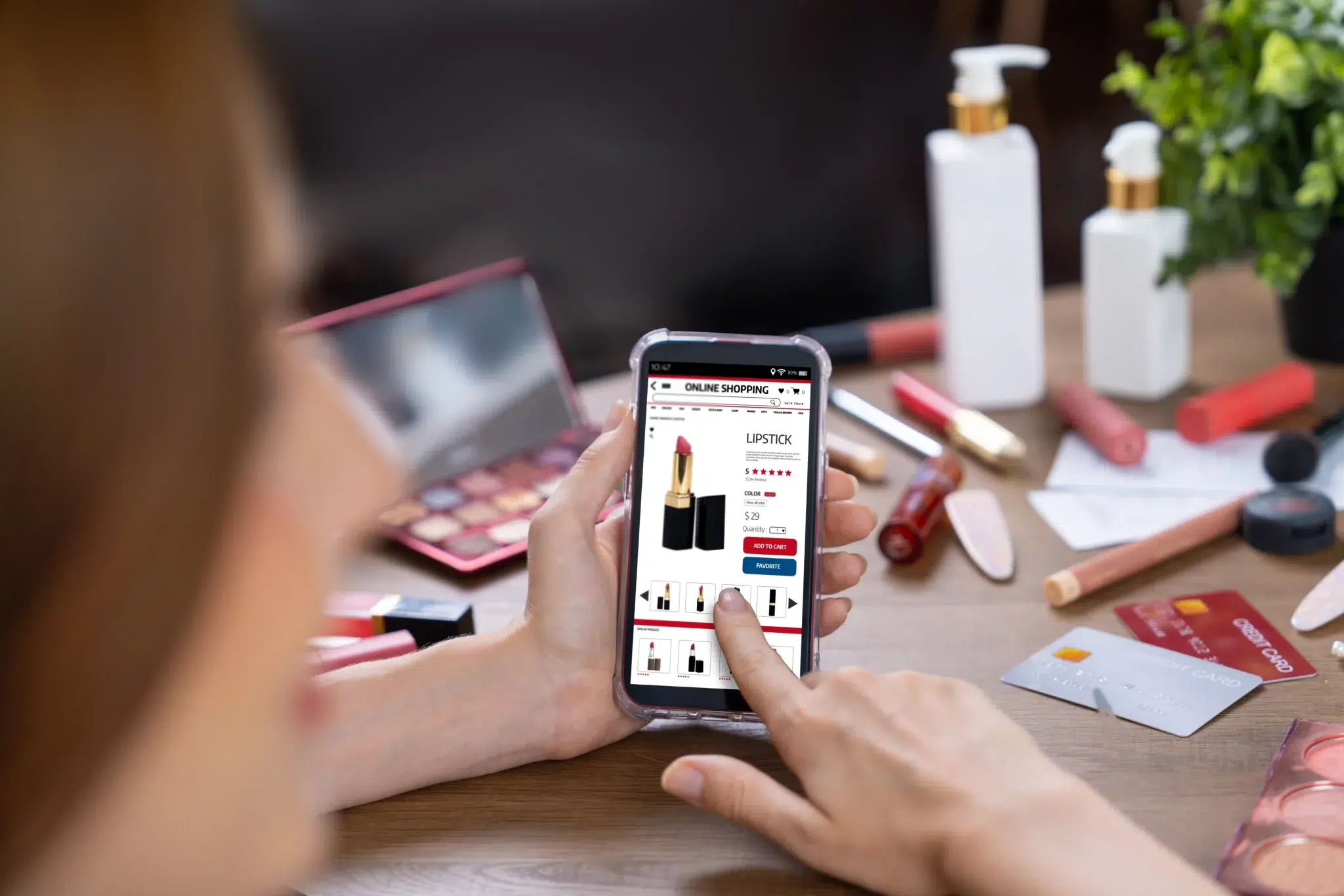 Person holding a smartphone showing an online lipstick product page amid makeup items on a table.