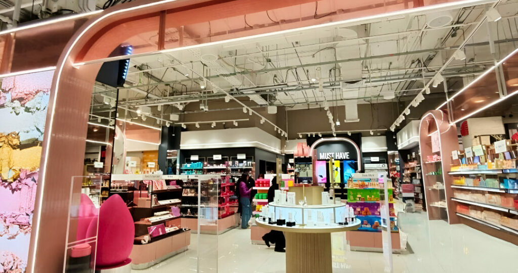 Bright cosmetics shop interior with a pink curved entrance, shelves of products, and a circular display table in the center as shoppers browse the aisles.