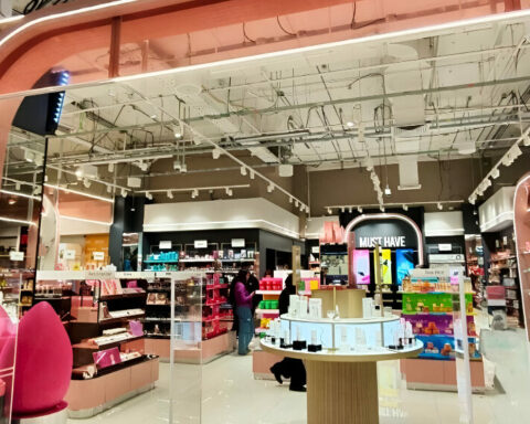 Bright cosmetics shop interior with a pink curved entrance, shelves of products, and a circular display table in the center as shoppers browse the aisles.