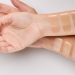 Multiple makeup foundation swatches in beige to tan tones along forearm for shade comparison on a light background.