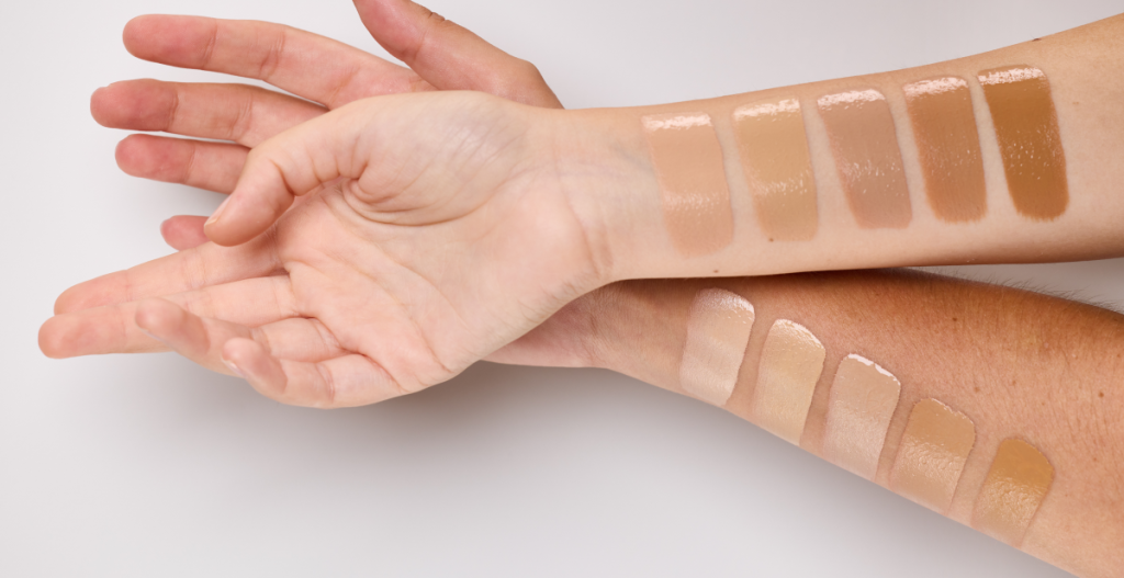 Multiple makeup foundation swatches in beige to tan tones along forearm for shade comparison on a light background.