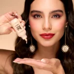 Close-up of a woman applying serum from a dropper bottle near her face, with bold makeup and statement earrings.