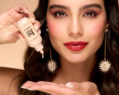 Close-up of a woman applying serum from a dropper bottle near her face, with bold makeup and statement earrings.