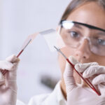 Scientist wearing safety goggles and gloves holds a curved glass sheet with tweezers in a lab setting.