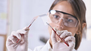 Scientist wearing safety goggles and gloves holds a curved glass sheet with tweezers in a lab setting.