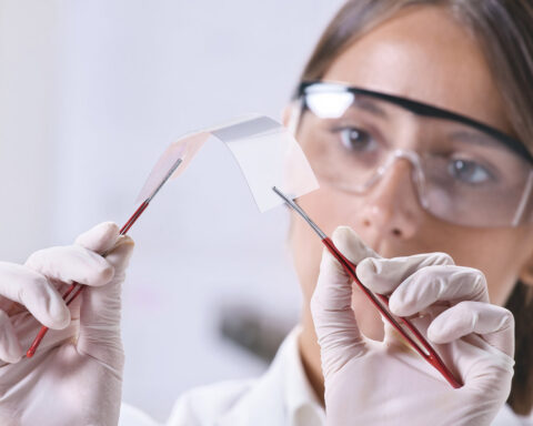 Scientist wearing safety goggles and gloves holds a curved glass sheet with tweezers in a lab setting.
