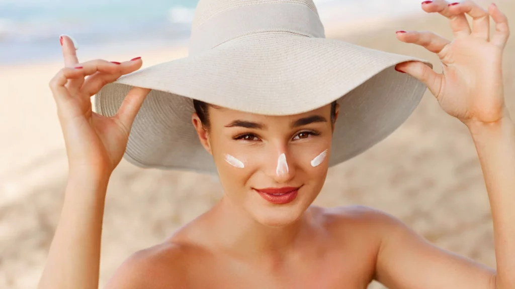 Woman at the beach wears a wide-brimmed hat and dots sunscreen on her face with visible white sunscreen on her nose and cheeks.