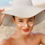 Woman at the beach wears a wide-brimmed hat and dots sunscreen on her face with visible white sunscreen on her nose and cheeks.