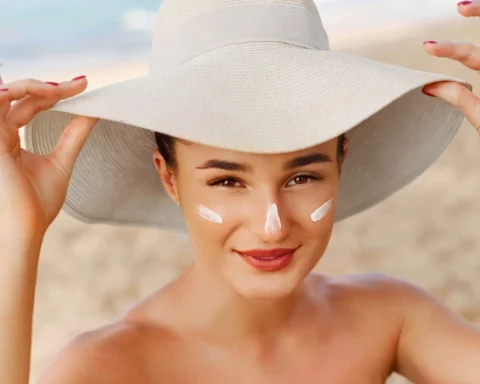 Woman at the beach wears a wide-brimmed hat and dots sunscreen on her face with visible white sunscreen on her nose and cheeks.